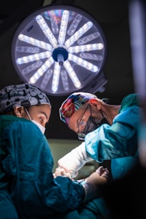 Two surgeons wearing masks and colorful caps are intensely focused on performing surgery under a bright surgical light. They are dressed in teal surgical gowns and white gloves, with a variety of surgical instruments in use.