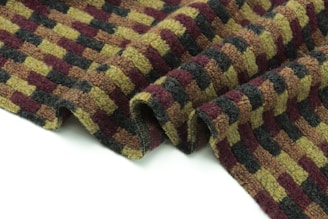 A textured fabric with a checkered pattern in alternating rows of olive green, burgundy, and dark gray. The material appears thick and woolen, with soft, woven ridges. It lies partially draped and slightly folded against a plain white background.