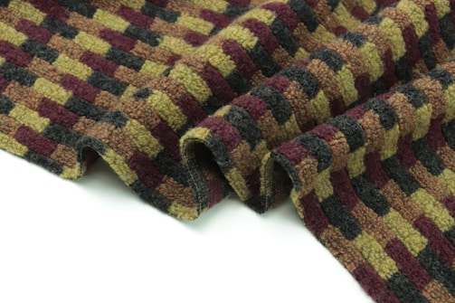 A textured fabric with a checkered pattern in alternating rows of olive green, burgundy, and dark gray. The material appears thick and woolen, with soft, woven ridges. It lies partially draped and slightly folded against a plain white background.