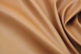 A close-up view of smooth, tan-colored leather fabric with gentle folds and a soft texture. The leather has a subtle sheen and fine grain, giving it an elegant appearance.