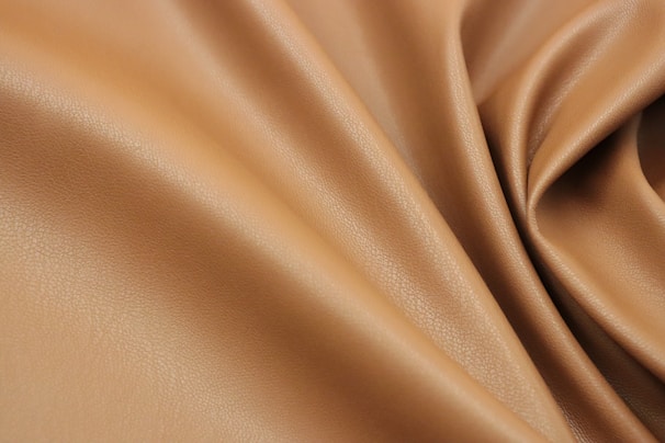 A close-up view of smooth, tan-colored leather fabric with gentle folds and a soft texture. The leather has a subtle sheen and fine grain, giving it an elegant appearance.
