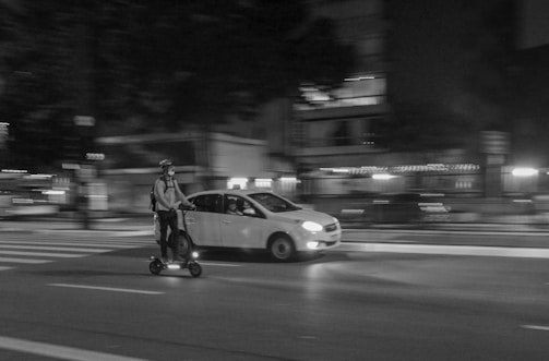 A nighttime urban scene where a person wearing a helmet and backpack rides an electric scooter on a city street. A car is driving alongside in the same direction, and blurred lights from nearby buildings suggest movement and speed.