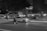 A nighttime urban scene where a person wearing a helmet and backpack rides an electric scooter on a city street. A car is driving alongside in the same direction, and blurred lights from nearby buildings suggest movement and speed.
