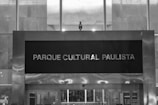 A black and white image featuring a large sign with the text 'Parque Cultural Paulista' mounted on a modern building facade. The design consists of clean, straight lines with a mixture of metal and glass materials. Light reflects softly off the metallic surfaces, creating a sleek and urban look.
