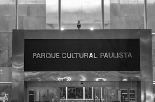 A black and white image featuring a large sign with the text 'Parque Cultural Paulista' mounted on a modern building facade. The design consists of clean, straight lines with a mixture of metal and glass materials. Light reflects softly off the metallic surfaces, creating a sleek and urban look.