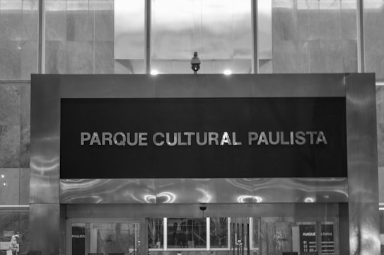 A black and white image featuring a large sign with the text 'Parque Cultural Paulista' mounted on a modern building facade. The design consists of clean, straight lines with a mixture of metal and glass materials. Light reflects softly off the metallic surfaces, creating a sleek and urban look.