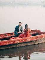 A man in a suit and a woman in a red dress are seated in a wooden boat on a calm body of water. The background is misty with a few birds flying in the distance, creating a serene and romantic atmosphere.