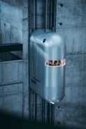 A futuristic, sleek elevator is positioned against a concrete wall. The carriage has a shiny, metallic exterior with a narrow window at the front, suggesting a modern or advanced design. The surrounding structure is predominantly built of raw, grey concrete, enhancing the industrial and modern aesthetic.
