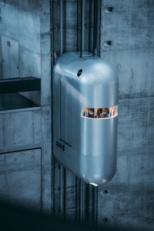 A futuristic, sleek elevator is positioned against a concrete wall. The carriage has a shiny, metallic exterior with a narrow window at the front, suggesting a modern or advanced design. The surrounding structure is predominantly built of raw, grey concrete, enhancing the industrial and modern aesthetic.