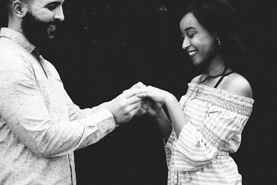 A black and white image depicting two people holding hands and smiling at each other. The man on the left has a beard and is wearing a light-colored, long-sleeved shirt. The woman on the right has long hair and wears an off-shoulder patterned top.