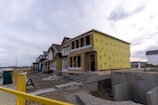 A residential construction site features several houses in various stages of development. The house in the foreground is partially built, with its structure framed and covered in yellow Tyvek sheathing. Other houses in the background appear more complete, displaying dark blue and mustard yellow siding. A yellow temporary fence runs along the edge of the site, and construction materials are scattered across the ground. A cloudy sky looms overhead, contributing to a subdued atmosphere.