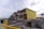 A residential construction site features several houses in various stages of development. The house in the foreground is partially built, with its structure framed and covered in yellow Tyvek sheathing. Other houses in the background appear more complete, displaying dark blue and mustard yellow siding. A yellow temporary fence runs along the edge of the site, and construction materials are scattered across the ground. A cloudy sky looms overhead, contributing to a subdued atmosphere.
