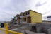 A residential construction site features several houses in various stages of development. The house in the foreground is partially built, with its structure framed and covered in yellow Tyvek sheathing. Other houses in the background appear more complete, displaying dark blue and mustard yellow siding. A yellow temporary fence runs along the edge of the site, and construction materials are scattered across the ground. A cloudy sky looms overhead, contributing to a subdued atmosphere.