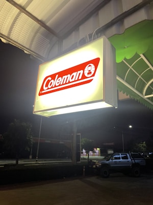 A brightly illuminated sign with the brand name 'Coleman' in red and white hangs under a canopy at night. Below, a parking area is visible with a parked vehicle to the right. The scene is set against a dark night sky, with some trees and other structures visible in the background.