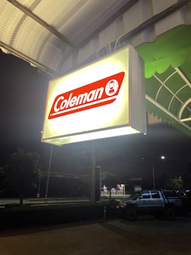 A brightly illuminated sign with the brand name 'Coleman' in red and white hangs under a canopy at night. Below, a parking area is visible with a parked vehicle to the right. The scene is set against a dark night sky, with some trees and other structures visible in the background.