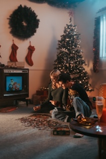 two people sitting on the floor in front of a christmas tree