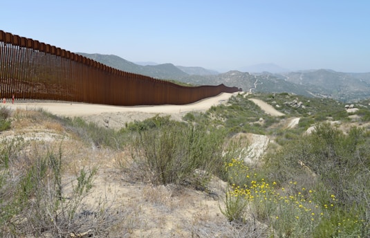 A long, rust-colored fence stretches across a hilly, arid terrain. The landscape is characterized by sparse vegetation, rocky ground, and patches of yellow wildflowers. The sky is clear blue, and distant mountains are visible, suggesting a border setting.