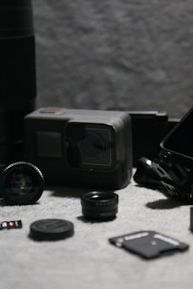 A collection of camera accessories including a GoPro camera, multiple lens caps, a microSD card, and various adapters arranged on a textured grey fabric surface. The image is composed with a shallow depth of field, highlighting the main objects while the background is blurred.