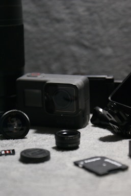 A collection of camera accessories including a GoPro camera, multiple lens caps, a microSD card, and various adapters arranged on a textured grey fabric surface. The image is composed with a shallow depth of field, highlighting the main objects while the background is blurred.