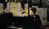 A person sits at a cluttered desk, surrounded by electronic equipment, books, and decorative artwork with intricate patterns depicting stylized creatures. The individual appears to be deep in thought or possibly stressed, covering their face partially with one hand. A warm desk lamp illuminates the space.