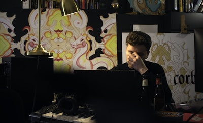 A person sits at a cluttered desk, surrounded by electronic equipment, books, and decorative artwork with intricate patterns depicting stylized creatures. The individual appears to be deep in thought or possibly stressed, covering their face partially with one hand. A warm desk lamp illuminates the space.