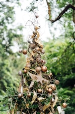A decorative outdoor ornament resembling a Christmas tree is adorned with golden ribbons, metallic gold and bronze baubles, and a large decorative star. The surrounding lush greenery enhances the festive look.