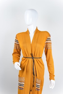 A mannequin is dressed in a mustard-yellow cardigan featuring ornate patterns on the shoulders and pockets. The cardigan is styled with a thin black belt around the waist, and the mannequin stands against a plain white background.