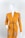 A mannequin is dressed in a mustard-yellow cardigan featuring ornate patterns on the shoulders and pockets. The cardigan is styled with a thin black belt around the waist, and the mannequin stands against a plain white background.