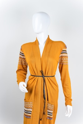 A mannequin is dressed in a mustard-yellow cardigan featuring ornate patterns on the shoulders and pockets. The cardigan is styled with a thin black belt around the waist, and the mannequin stands against a plain white background.