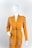 A mannequin is dressed in a mustard-yellow cardigan featuring ornate patterns on the shoulders and pockets. The cardigan is styled with a thin black belt around the waist, and the mannequin stands against a plain white background.