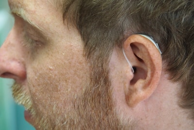 A close-up view of a person's ear with a hearing aid fitted behind it. The image focuses on the side profile, showing facial hair and skin texture.