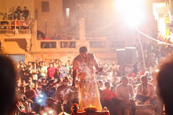 A large crowd gathers in an outdoor urban setting where a man is performing a ceremony or ritual with smoke emanating from an object in front of him. The scene is brightly lit, possibly indicating a nighttime event, with spectators watching attentively.