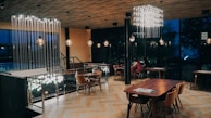 A modern, elegantly designed dining area with wooden flooring and a variety of lighting fixtures. The space features wooden tables and chairs arranged neatly, with subtle lighting casting a warm ambiance. The walls are mostly glass, revealing a dark, outdoor view through the windows.