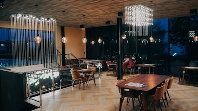 A modern, elegantly designed dining area with wooden flooring and a variety of lighting fixtures. The space features wooden tables and chairs arranged neatly, with subtle lighting casting a warm ambiance. The walls are mostly glass, revealing a dark, outdoor view through the windows.