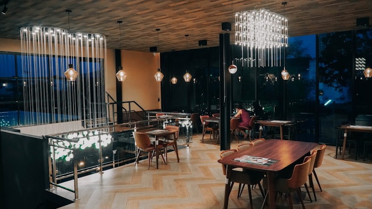 A modern, elegantly designed dining area with wooden flooring and a variety of lighting fixtures. The space features wooden tables and chairs arranged neatly, with subtle lighting casting a warm ambiance. The walls are mostly glass, revealing a dark, outdoor view through the windows.