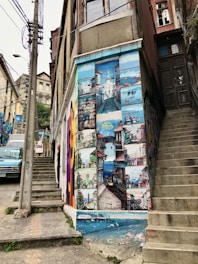 A mural painted on the side of a building, depicting various colorful scenes of a coastal town with boats, buildings, and a lighthouse. The mural is vibrant and detailed, spanning the height of the building wall. Adjacent are concrete stairs and an old door, with a person partially visible on a smaller staircase.