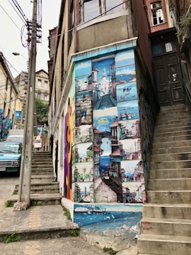 A mural painted on the side of a building, depicting various colorful scenes of a coastal town with boats, buildings, and a lighthouse. The mural is vibrant and detailed, spanning the height of the building wall. Adjacent are concrete stairs and an old door, with a person partially visible on a smaller staircase.
