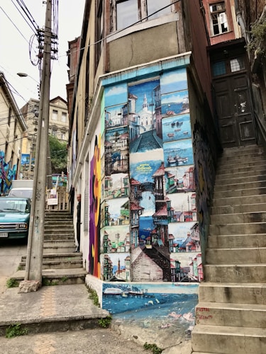 A mural painted on the side of a building, depicting various colorful scenes of a coastal town with boats, buildings, and a lighthouse. The mural is vibrant and detailed, spanning the height of the building wall. Adjacent are concrete stairs and an old door, with a person partially visible on a smaller staircase.