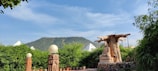 A peaceful outdoor scene with a large Buddha statue under a sprawling artificial tree. The foreground features lush green plants in pots, and decorative stone lamps with spherical lights. In the background, tents and a green hill are visible under a clear blue sky.