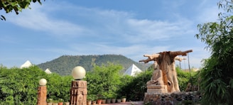 A peaceful outdoor scene with a large Buddha statue under a sprawling artificial tree. The foreground features lush green plants in pots, and decorative stone lamps with spherical lights. In the background, tents and a green hill are visible under a clear blue sky.