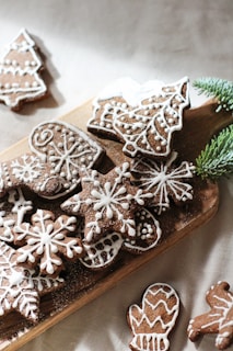 An assortment of brown gingerbread cookies decorated with intricate white icing patterns, arranged on a wooden board. The shapes include snowflakes, mittens, trees, and other festive designs. A sprig of green pine needles adds a touch of natural decoration.