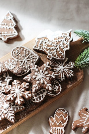 An assortment of brown gingerbread cookies decorated with intricate white icing patterns, arranged on a wooden board. The shapes include snowflakes, mittens, trees, and other festive designs. A sprig of green pine needles adds a touch of natural decoration.