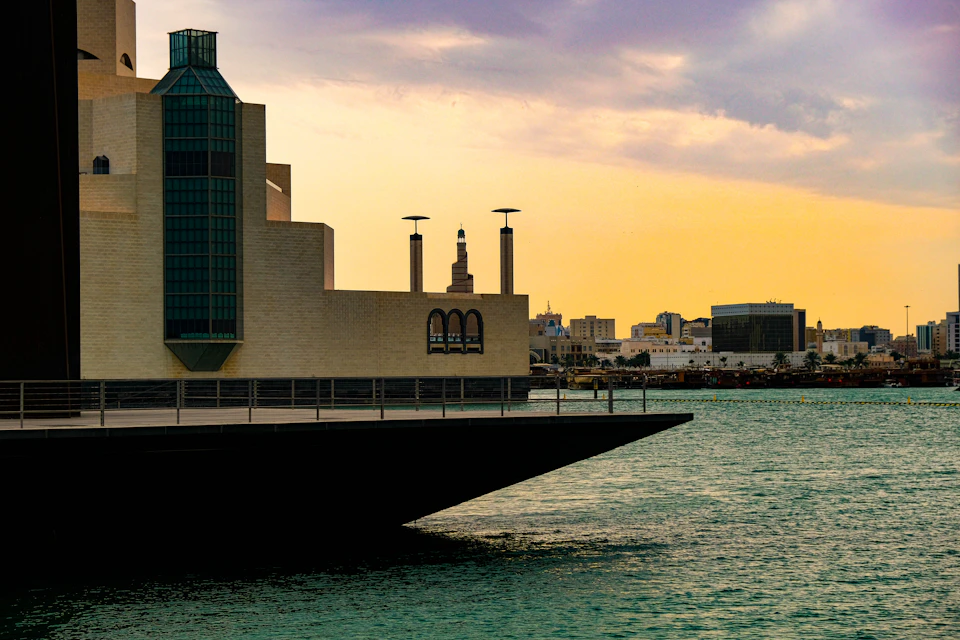 The image depicts a serene cityscape at sunset, with a large building featuring a green roof and two towers on its left side. The sky is painted in warm hues of orange and pink, creating a beautiful backdrop for the scene. The calm water below mirrors the colors of the sky, adding to the overall tranquility of the image. In the foreground, there are several people enjoying their time near the waterfront, some standing on a pier that extends into the water.