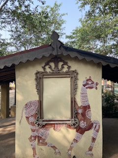 A decorative wall features a painted camel adorned with intricate white patterns. The center of the wall holds an ornate, empty picture frame with elaborate carvings, set against a backdrop of trees and a simple building structure.