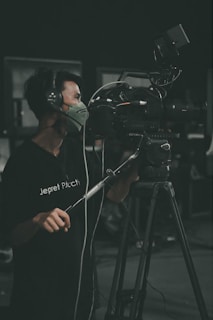 A person is operating a professional video camera on a tripod. They are wearing headphones and a face mask while focusing on filming. The setting appears to be in a dark room or studio.