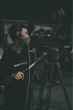 A person is operating a professional video camera on a tripod. They are wearing headphones and a face mask while focusing on filming. The setting appears to be in a dark room or studio.
