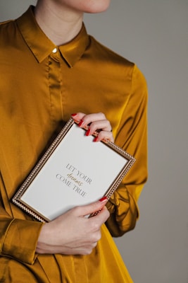 A person wearing a mustard yellow shirt is holding a framed message. The frame is gold and contains the text 'Let Your Dreams Come True'. The person's nails are painted red, and they are holding the frame gently with both hands against a plain background.