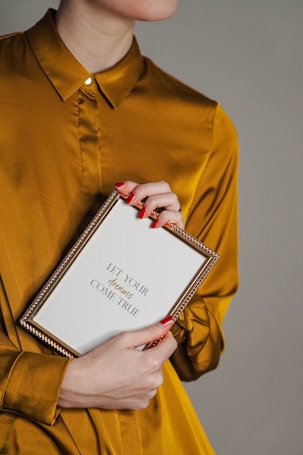 A person wearing a mustard yellow shirt is holding a framed message. The frame is gold and contains the text 'Let Your Dreams Come True'. The person's nails are painted red, and they are holding the frame gently with both hands against a plain background.
