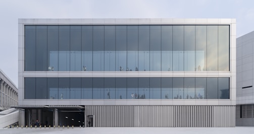 A modern building with a clean, minimalist design features large glass windows across three floors. The transparent facade allows a view of people moving within the building. There is a section at the bottom with metal cladding and an entrance area.