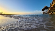 Golden light from the setting sun illuminates the shoreline, creating a serene interplay of colors. Waves gently lap at the sandy beach, while a wooden hut with thatched roofing extends over the water. The horizon is dotted with silhouettes of palm trees, conveying a peaceful, tropical atmosphere.